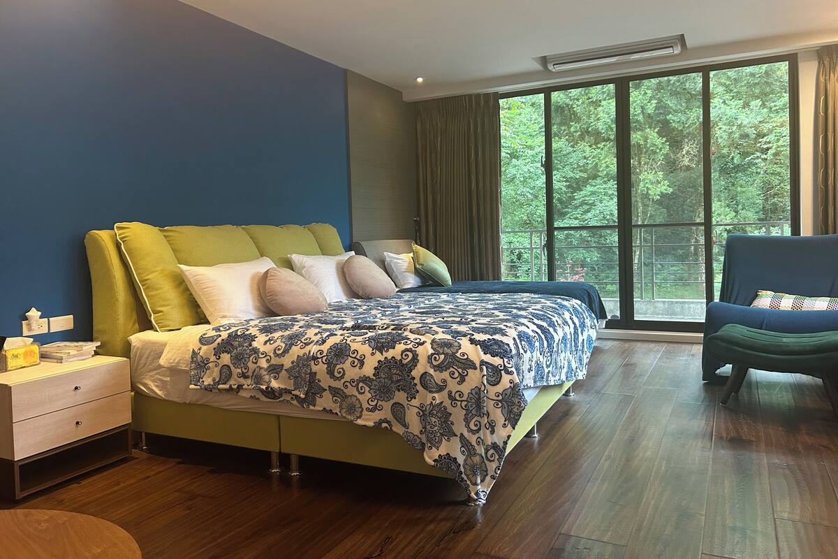 Un dormitorio con una cama grande, una mesita de noche, una silla y una ventana con vistas a árboles.