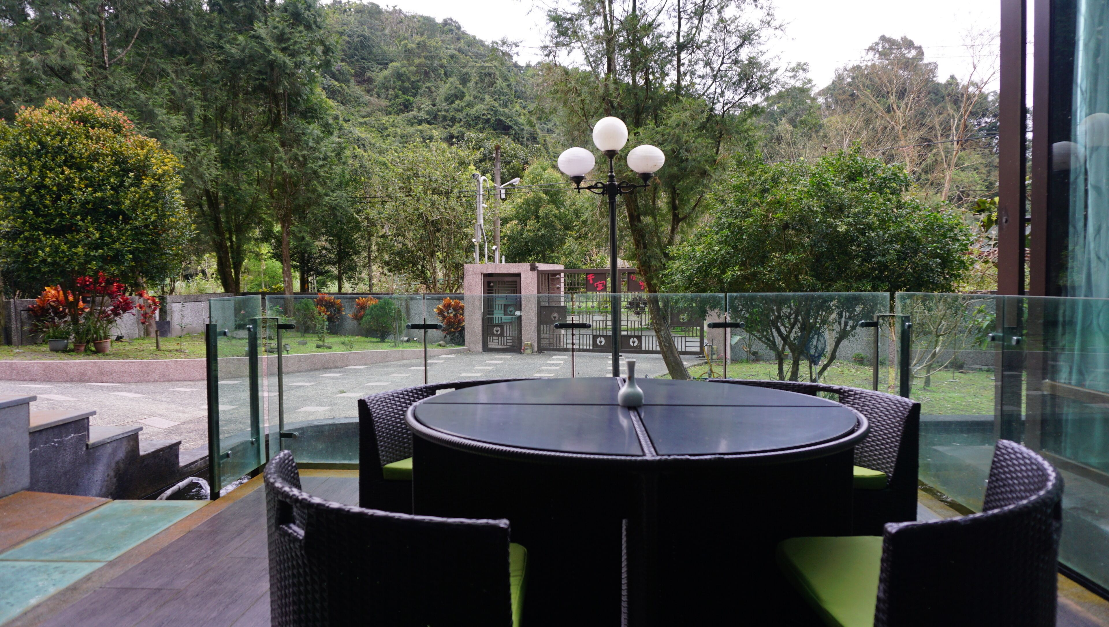 A round table with chairs in a glass-enclosed outdoor area, overlooking a garden with trees and a gate.