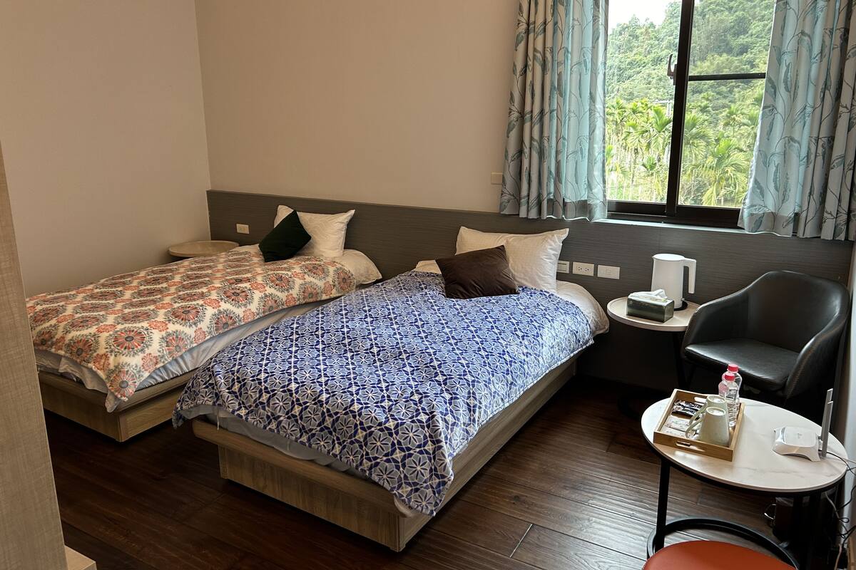 Habitación de hotel con dos camas, una mesita con hervidor de agua, una silla y vistas a zonas verdes.