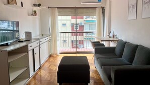 A modern living room with a grey sofa, a black ottoman, and a TV on a white cabinet. There is a balcony with a view of buildings outside.
