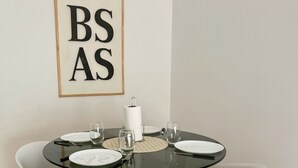 A round dining table with white chairs, a glass top, and a wooden base. There are plates, glasses, and a napkin holder on the table. A framed sign with the letters "BSAS" hangs on the wall above the table.
