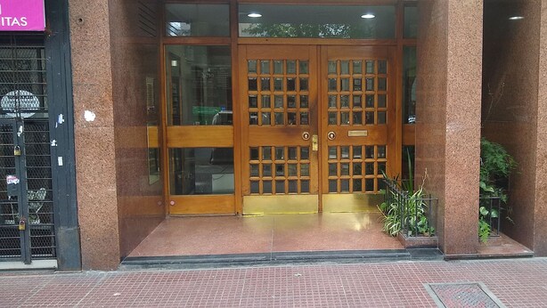 A building entrance with a double wooden door, a metal gate on the left, and a small potted plant on the right.