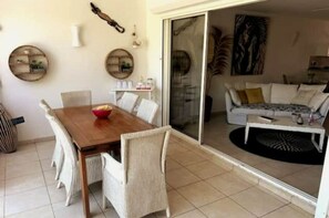 Un coin repas avec une table en bois et des chaises blanches, un salon avec un canapé et une table basse, et une porte-fenêtre coulissante donnant sur un balcon.