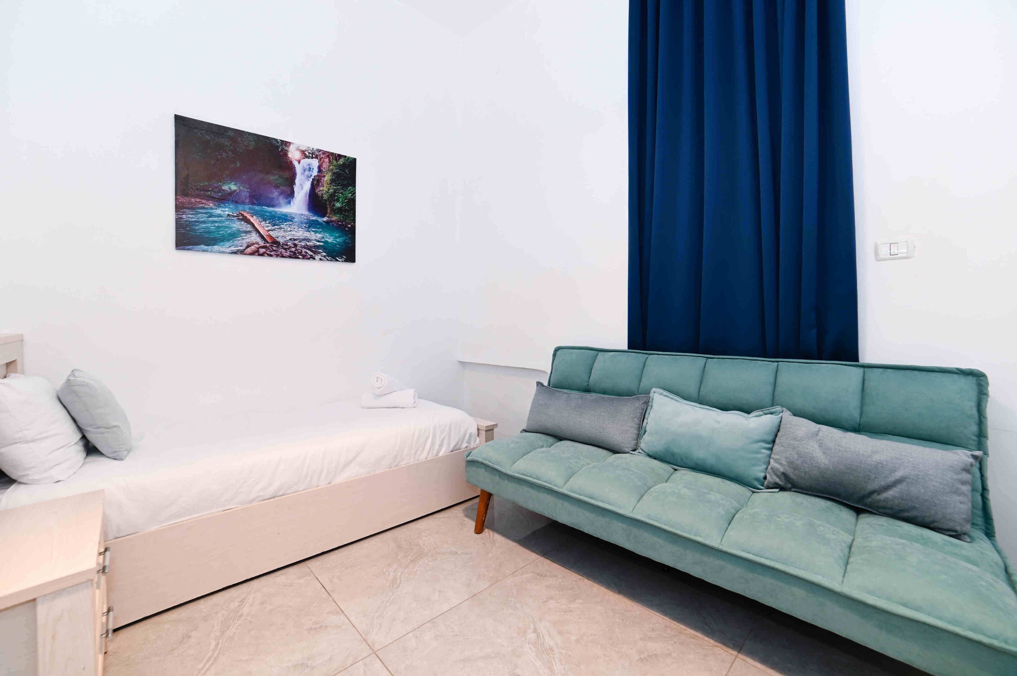 A room with a bed, a green sofa, a wall-mounted picture of a waterfall, and a blue curtain.