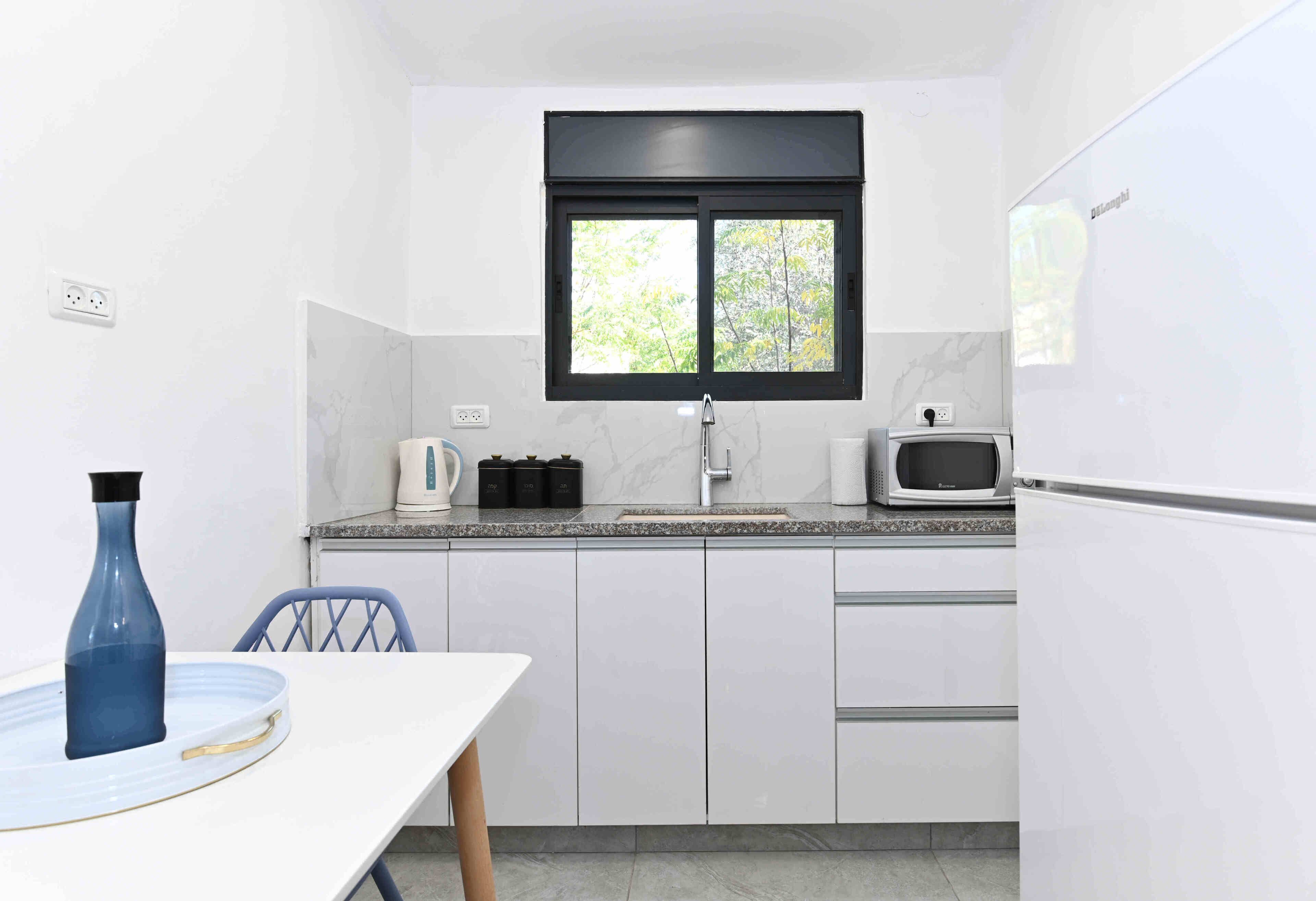 A compact kitchen with white cabinets, a microwave, a kettle, and a window above the sink.