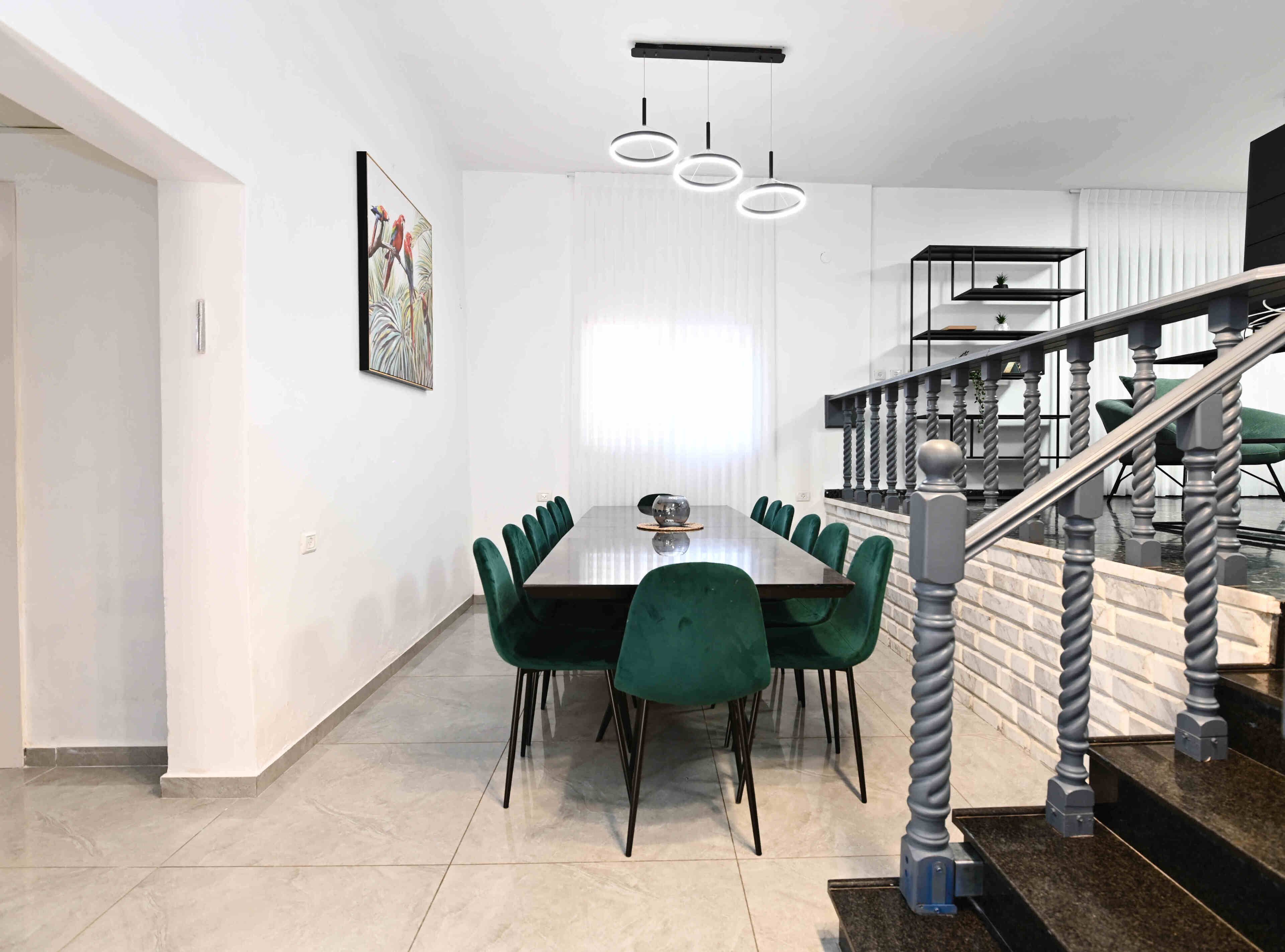 A modern dining area with a long table and green upholstered chairs, a staircase with a unique balustrade, and a framed artwork on the wall.