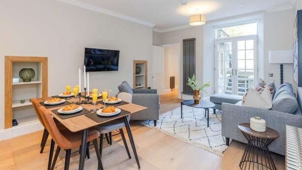 Ein modernes Wohnzimmer mit Essbereich, grauen Sofas, einem Fernseher und einem großen Fenster.