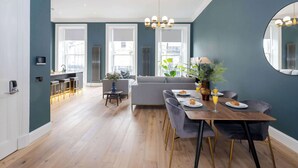 A modern living room with a dining area, large windows, and a kitchen in the background.