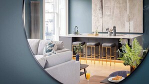 A modern living room with a sofa, a dining table with a glass of orange juice, and a kitchen area with stools and a large abstract painting.