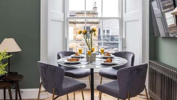 A dining area with a round table, four chairs, a vase with flowers, and a window with a view of buildings. - Jade Suite - City Centre 2 Bed Luxury Apartment (Edinburgh)
