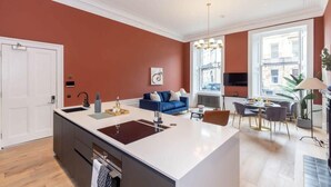 A modern kitchen with a central island, a dining area with a chandelier, and a blue sofa in a room with red walls. - Ember Suite - City Centre 1 Bed Luxury Apartment (Edinburgh)