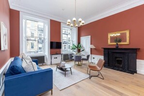 A living room with a blue sofa, a fireplace, a dining table with chairs, and large windows.