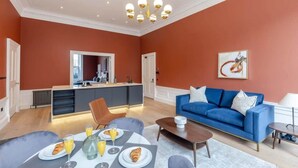 A modern living room with a blue sofa, a dining table with breakfast, and a kitchen area. - Ember Suite - City Centre 1 Bed Luxury Apartment (Edinburgh)
