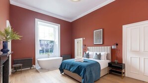 A bedroom with a large window, a bathtub, a bed with pillows, a bedside table, and a radiator. - Ember Suite - City Centre 1 Bed Luxury Apartment (Edinburgh)