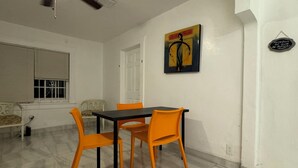 Ein Essbereich mit Tisch und vier orangefarbenen Stühlen, Deckenventilator, Fenster mit Jalousien und einem Wandbild.
