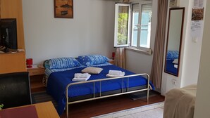 Una habitación de hotel con una cama individual, un escritorio con silla, un televisor sobre un mueble de madera, una ventana con cortinas y un espejo en la pared.