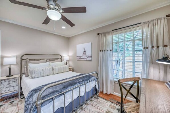 A bedroom with a bed, a ceiling fan, a window with curtains, a nightstand, and a chair.