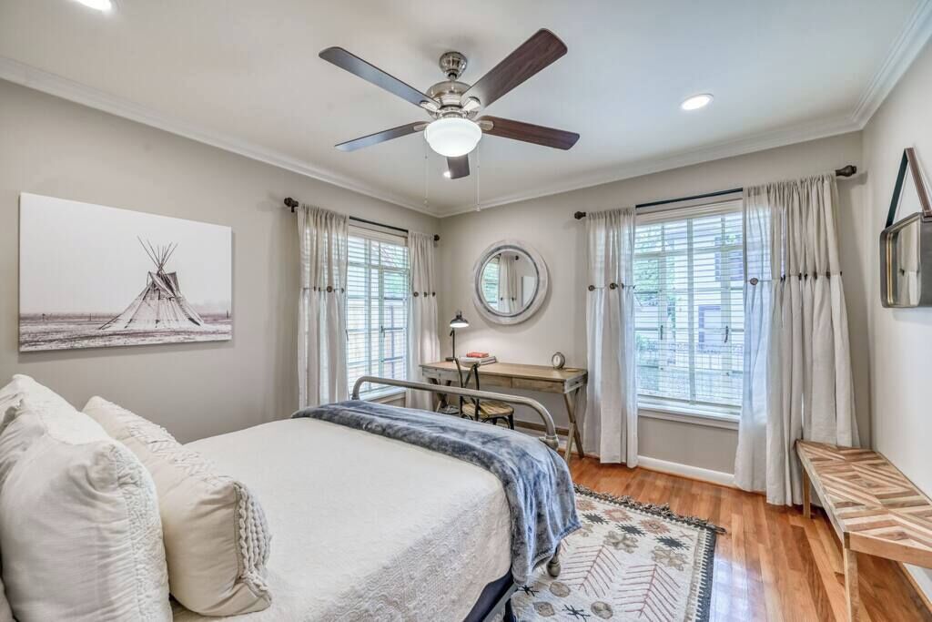 Ein Schlafzimmer mit einem Bett, einem Schreibtisch, einem Sessel, einem Deckenventilator, einem Fenster mit Vorhängen und einem Wandspiegel.