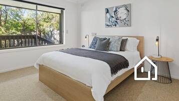 A bedroom with a large window, a bed with a wooden frame, a bedside table with a lamp, and a wall-mounted artwork.