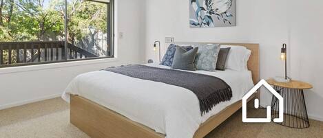 A bedroom with a large window, a bed with a wooden frame, a bedside table with a lamp, and a wall-mounted artwork.