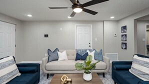 Un salon avec un canapé beige, deux fauteuils bleus, une table basse en bois, un ventilateur de plafond et une plante en pot.