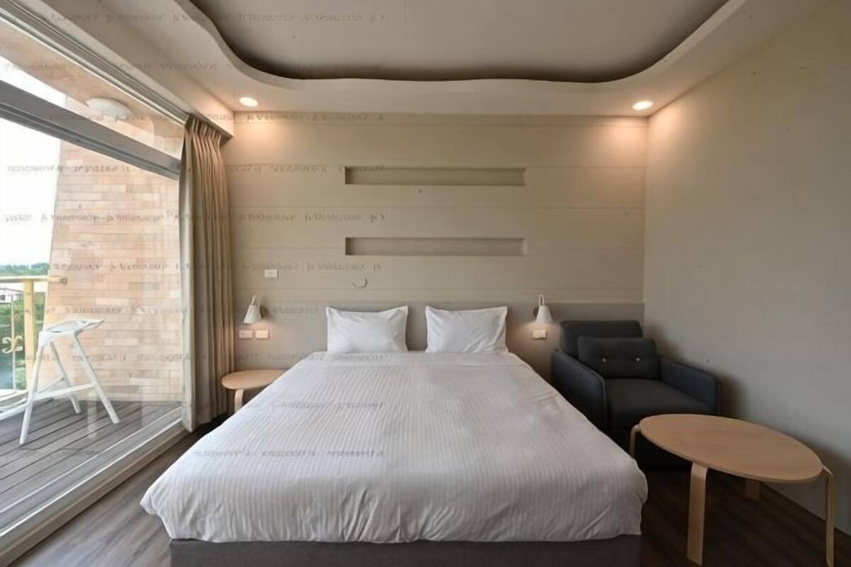Een moderne hotelkamer met een groot bed, twee nachtkastjes, een kleine zithoek en uitzicht op een balkon.