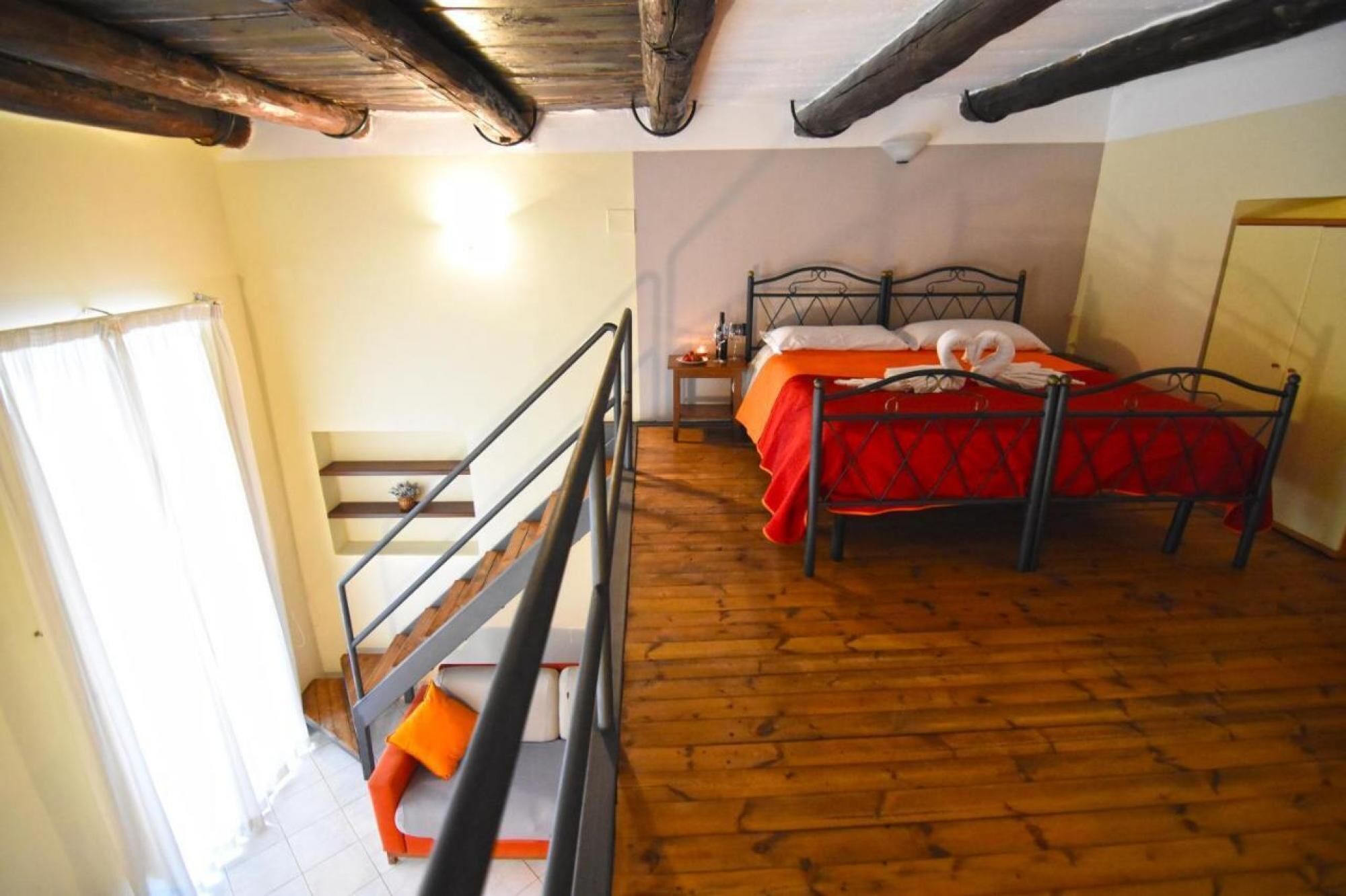 A room with a wooden floor, a bed with a red bedspread, a metal-framed headboard, and a staircase with a metal railing.