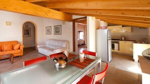 A cozy living room with a sofa, a dining table with chairs, a kitchen area with a refrigerator and stove, and wooden beams on the ceiling.