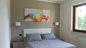 Une chambre à coucher comprenant un lit, deux tables de chevet, un tableau abstrait accroché au mur et une fenêtre donnant sur un paysage verdoyant.