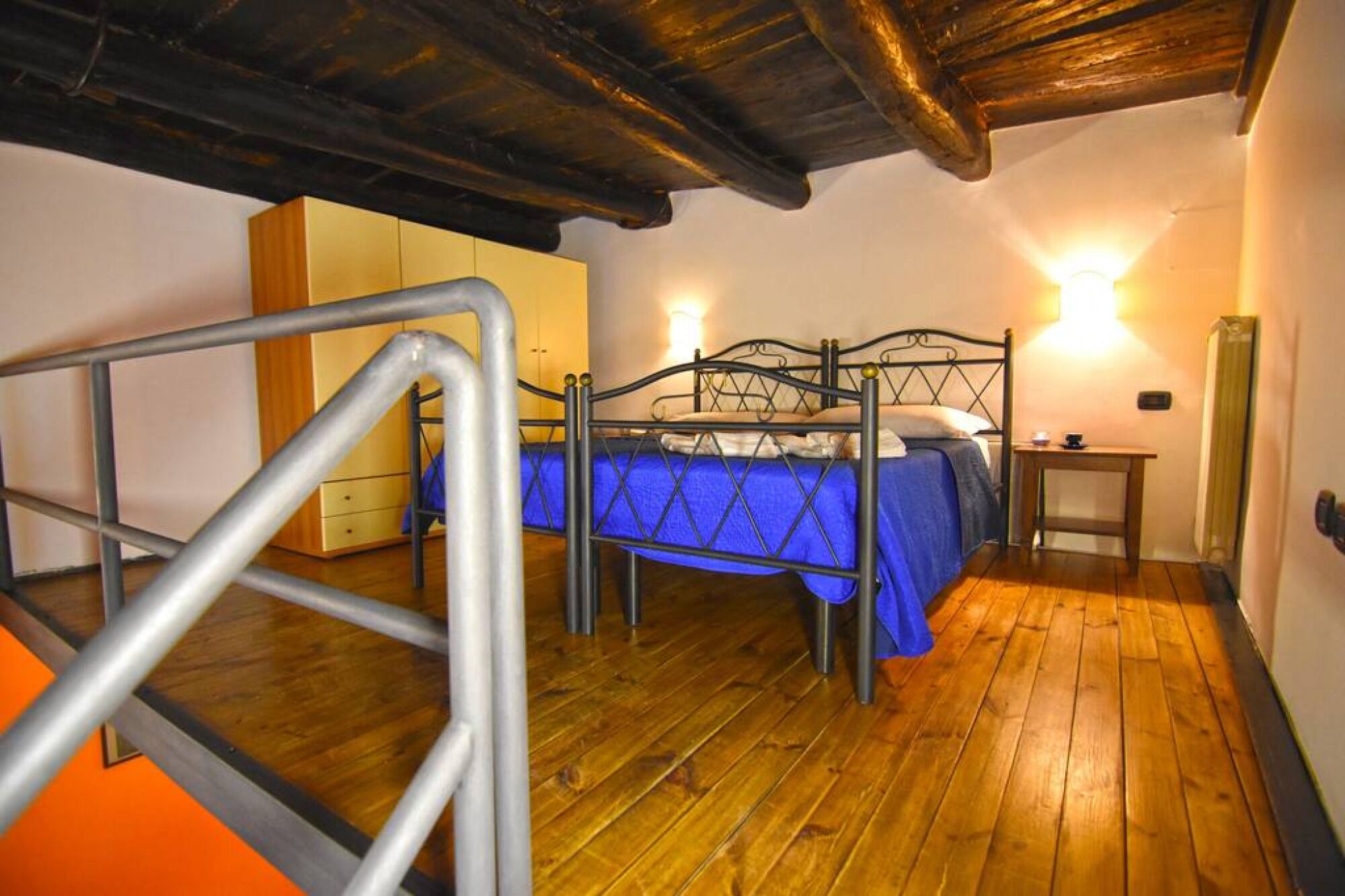 A bedroom with a metal bed frame, a blue bedspread, a bedside table, and a wall-mounted light.
