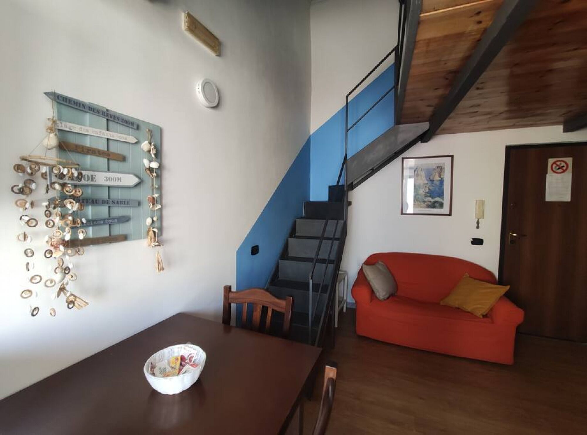Una zona de menjador amb una taula i cadires de fusta, un sofà vermell i una escala que condueix a un nivell superior.