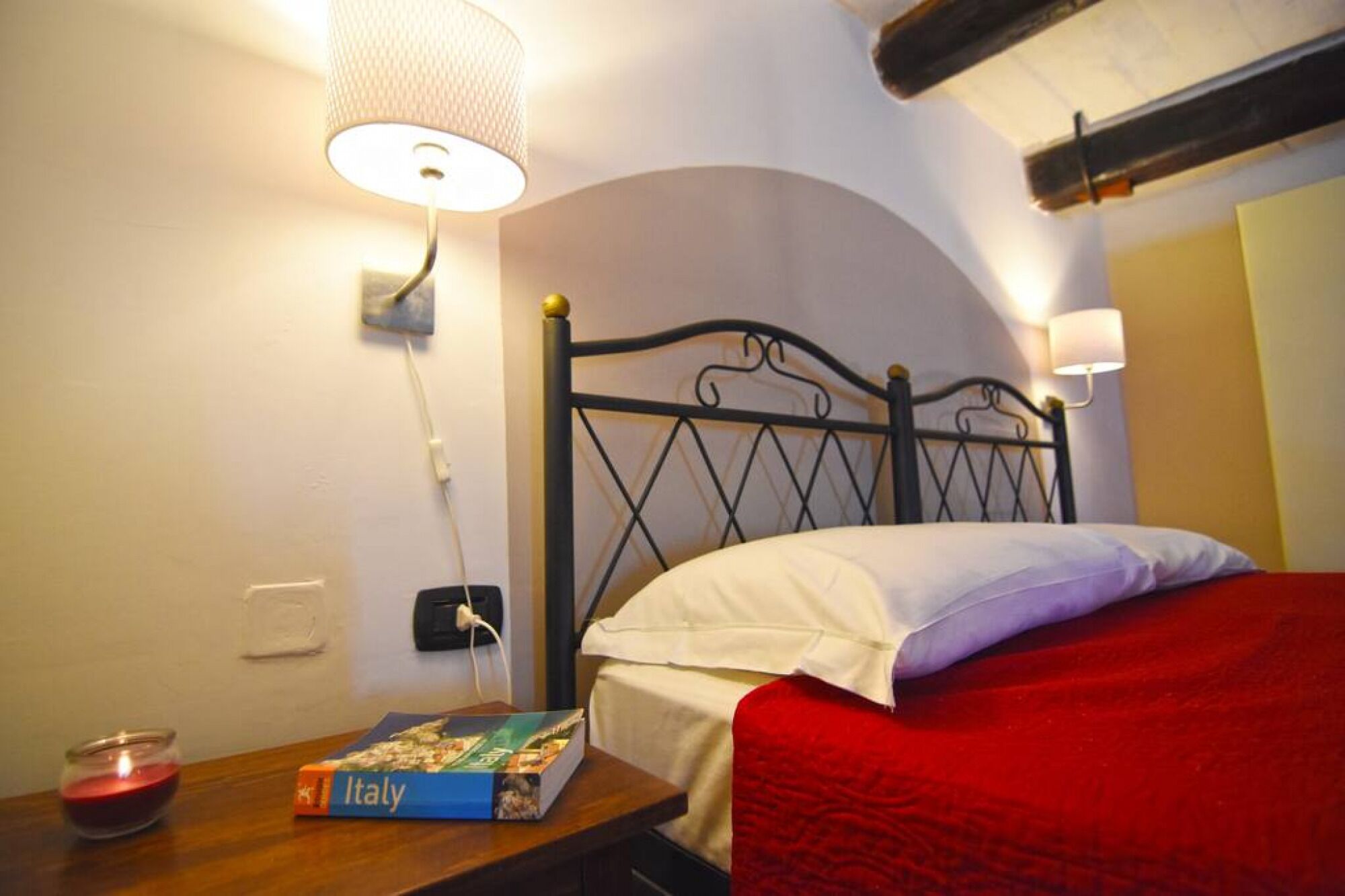 Un lit bien fait, recouvert d’une couverture rouge, une table de chevet sur laquelle repose un livre intitulé « Italie », et deux lampes fixées au mur.