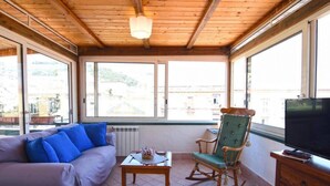 A cozy room with a wooden ceiling, a sofa with blue cushions, a rocking chair, a small table with a vase, and a television on a wooden cabinet. - Case Cosi Napoli - APP 17 (Naples)