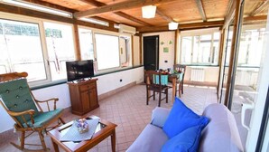 Een knus ingerichte kamer met een houten plafond, een bank met blauwe kussens, een schommelstoel, een televisie op een houten kast en een eettafel met stoelen.