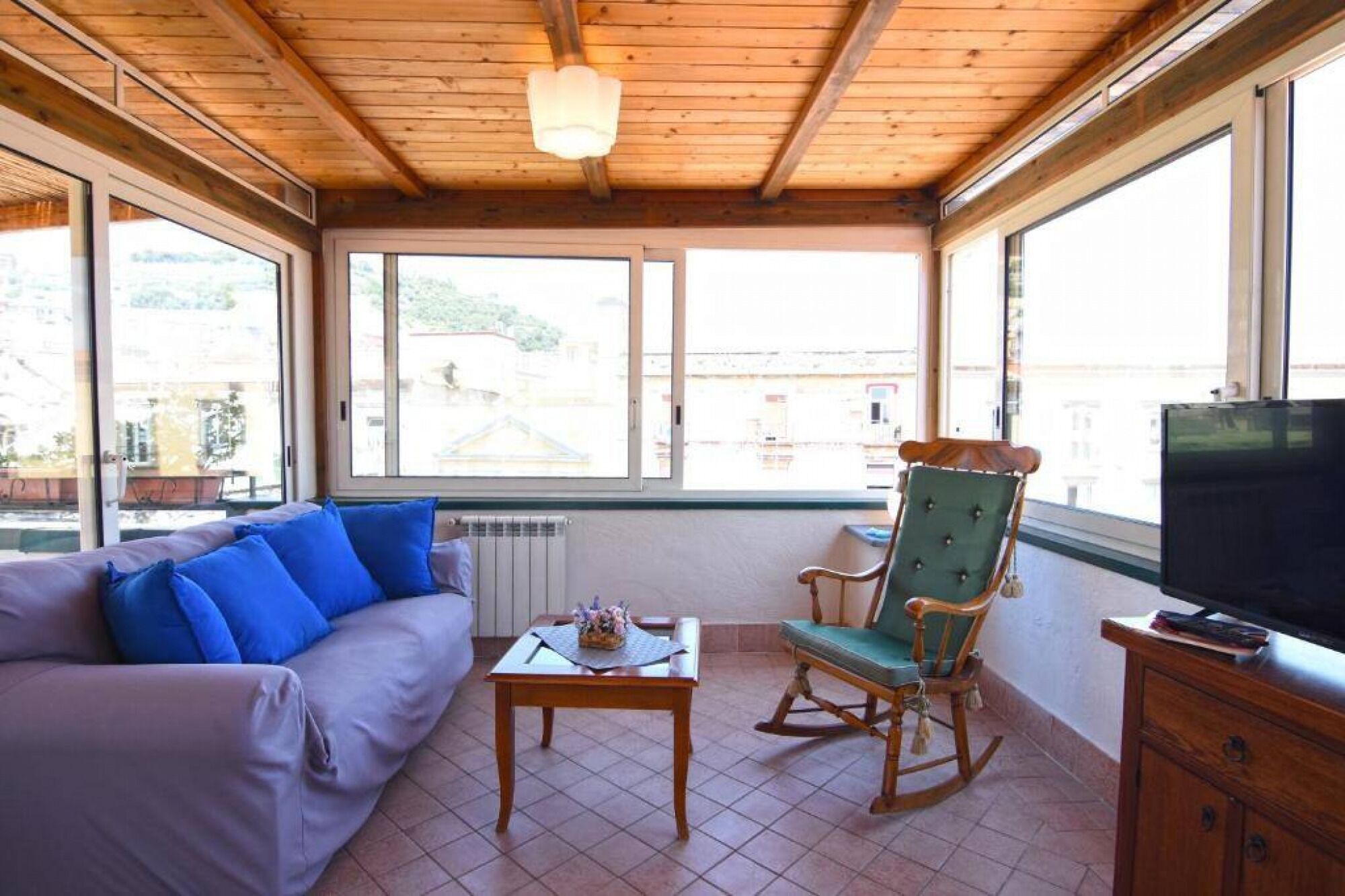 A cozy room with a wooden ceiling, a sofa with blue cushions, a rocking chair, a small table with a vase, and a television on a wooden cabinet.