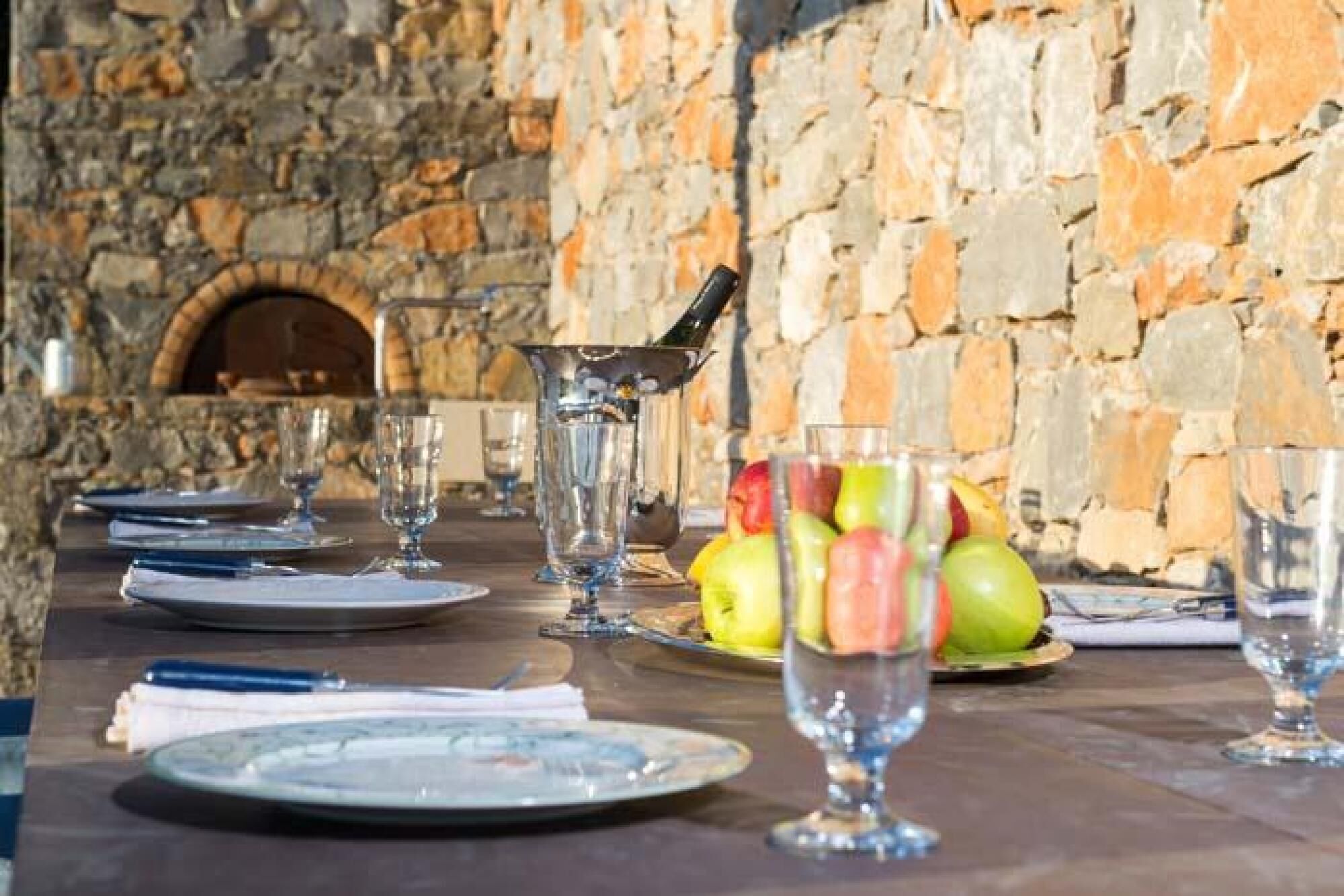 Un mur en pierre avec une cheminée, une table dressée avec des assiettes, des verres et un panier de fruits.