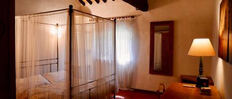 A four-poster bed with sheer curtains, a wooden nightstand with a lamp, and a mirror on the wall.