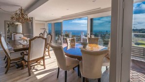 Un comedor con una mesa redonda y sillas, ventanas amplias con vista al mar y una araña.