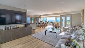 Amplia sala de estar con televisor de pantalla plana grande, un conjunto de sofás, zona de comedor con mesa y sillas, y balcón con vistas.