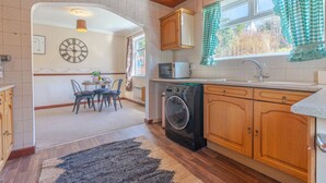 A kitchen with wooden cabinets, a washing machine, and a dining area with chairs and a table. - Melrose 2 (Lancaster)