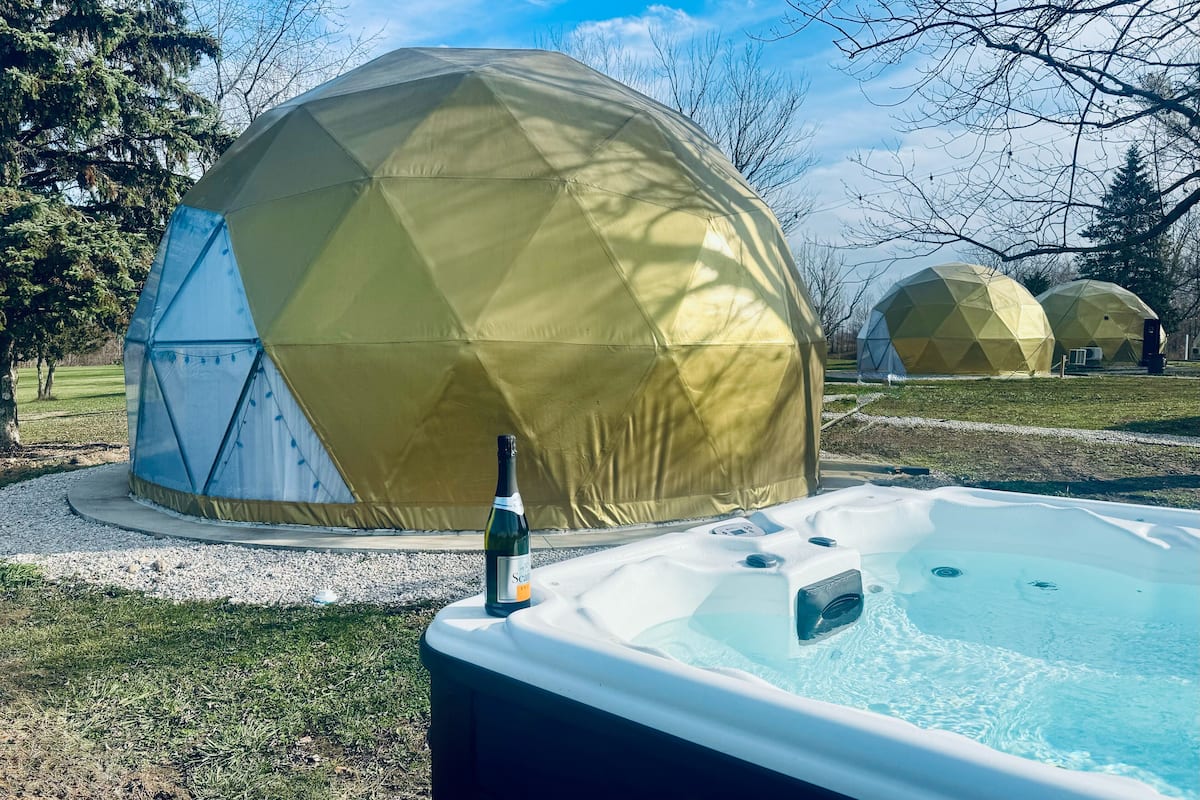 A hot tub with a bottle of wine, surrounded by a garden with a large geodesic dome structure.