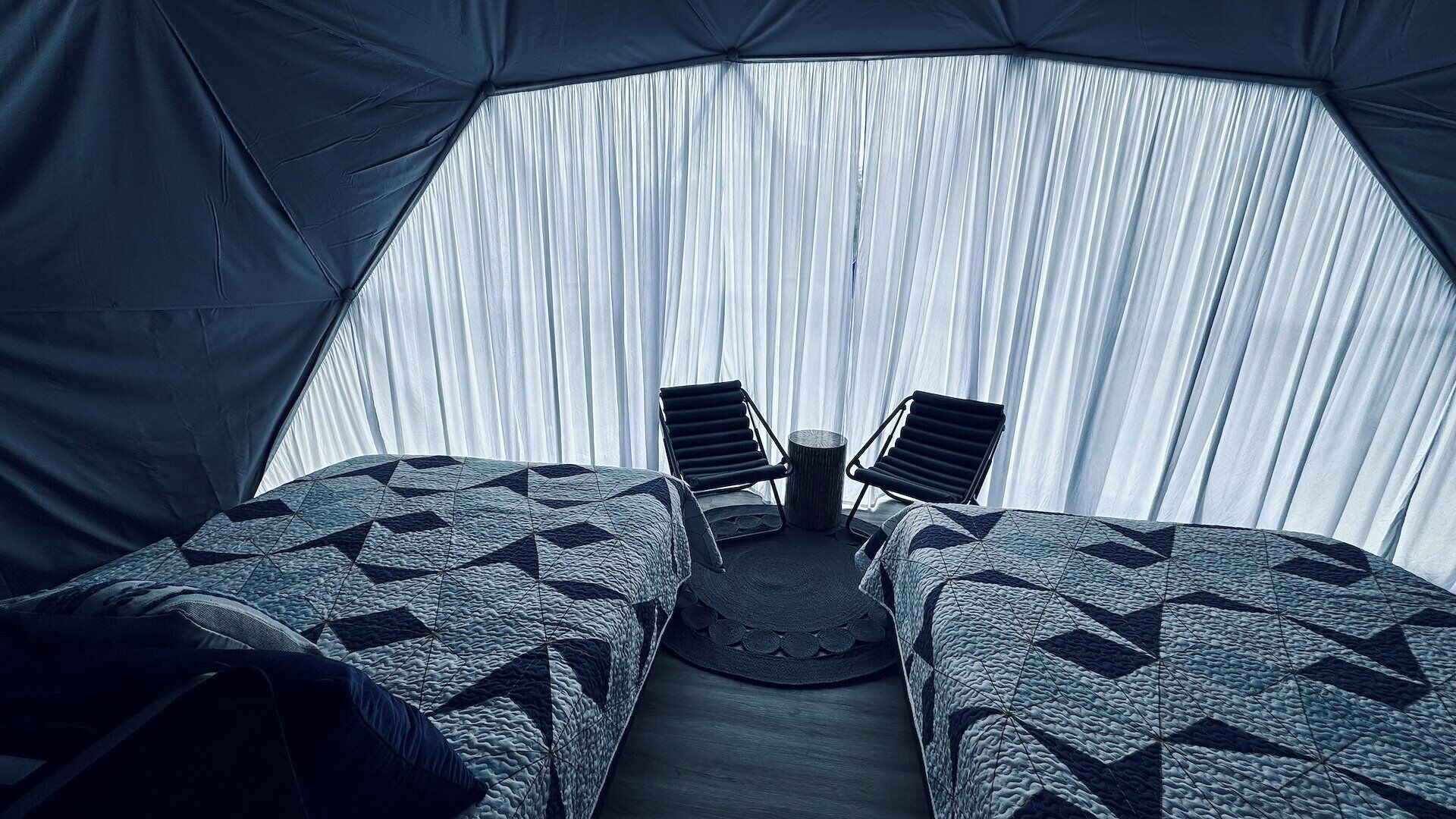 Eine Zelthotelunterkunft mit zwei Betten, jeweils mit einem geometrisch gemusterten Bettbezug, und einem kleinen runden Tisch dazwischen.