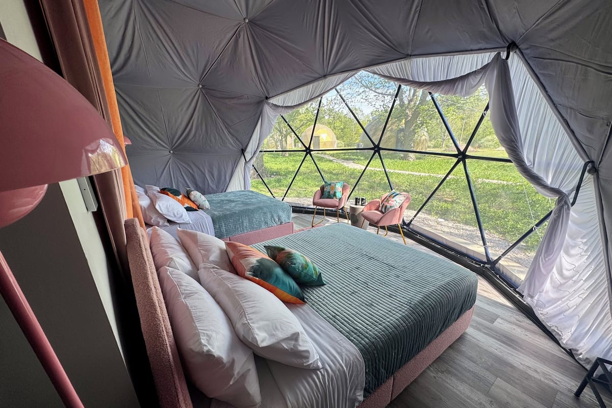 A spacious tented accommodation with two beds, each with multiple pillows and a green blanket. There are two chairs and a small table with a bottle and a cup. The tent has a large window offering a view of greenery outside.