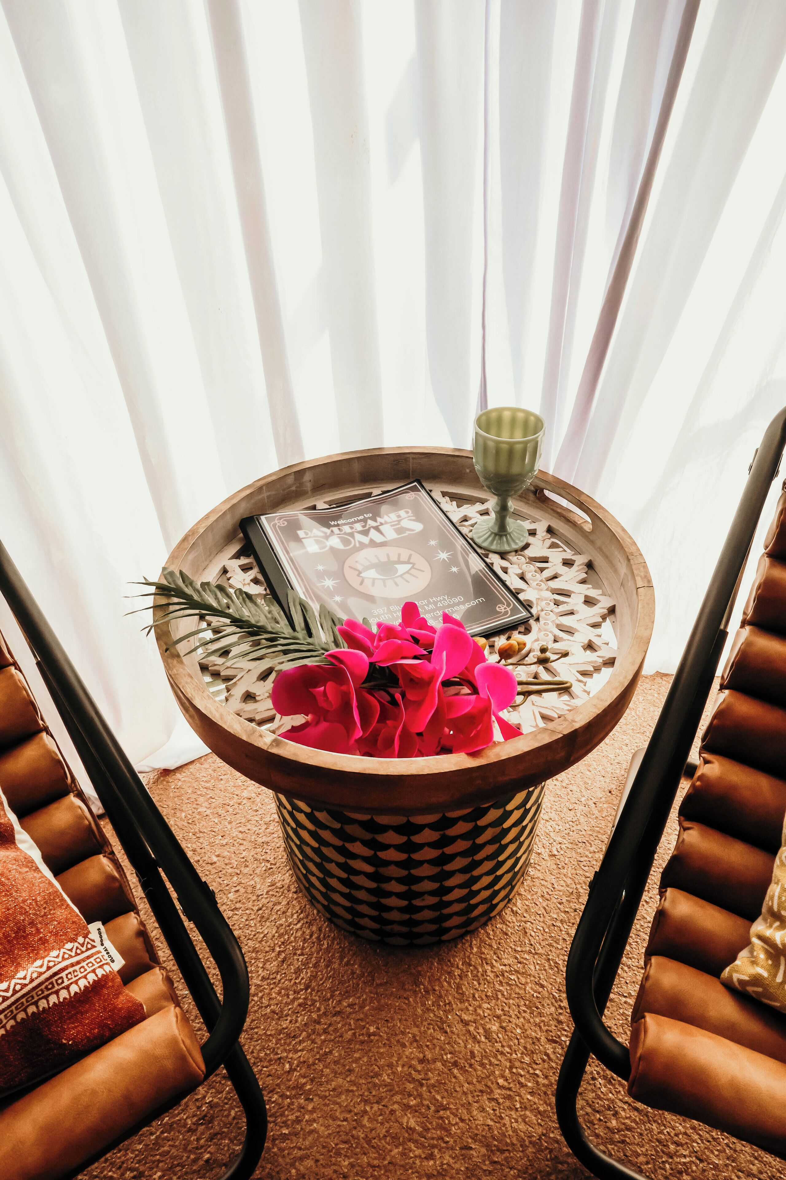 A round wooden table with a decorative tray, a book, a green glass, and a vase of flowers.