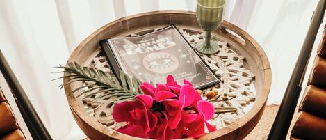 A round wooden table with a decorative tray, a book, a green glass, and a vase of flowers.