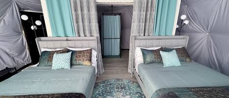 A room with two beds, grey upholstered headboards, patterned curtains, and a patterned rug.