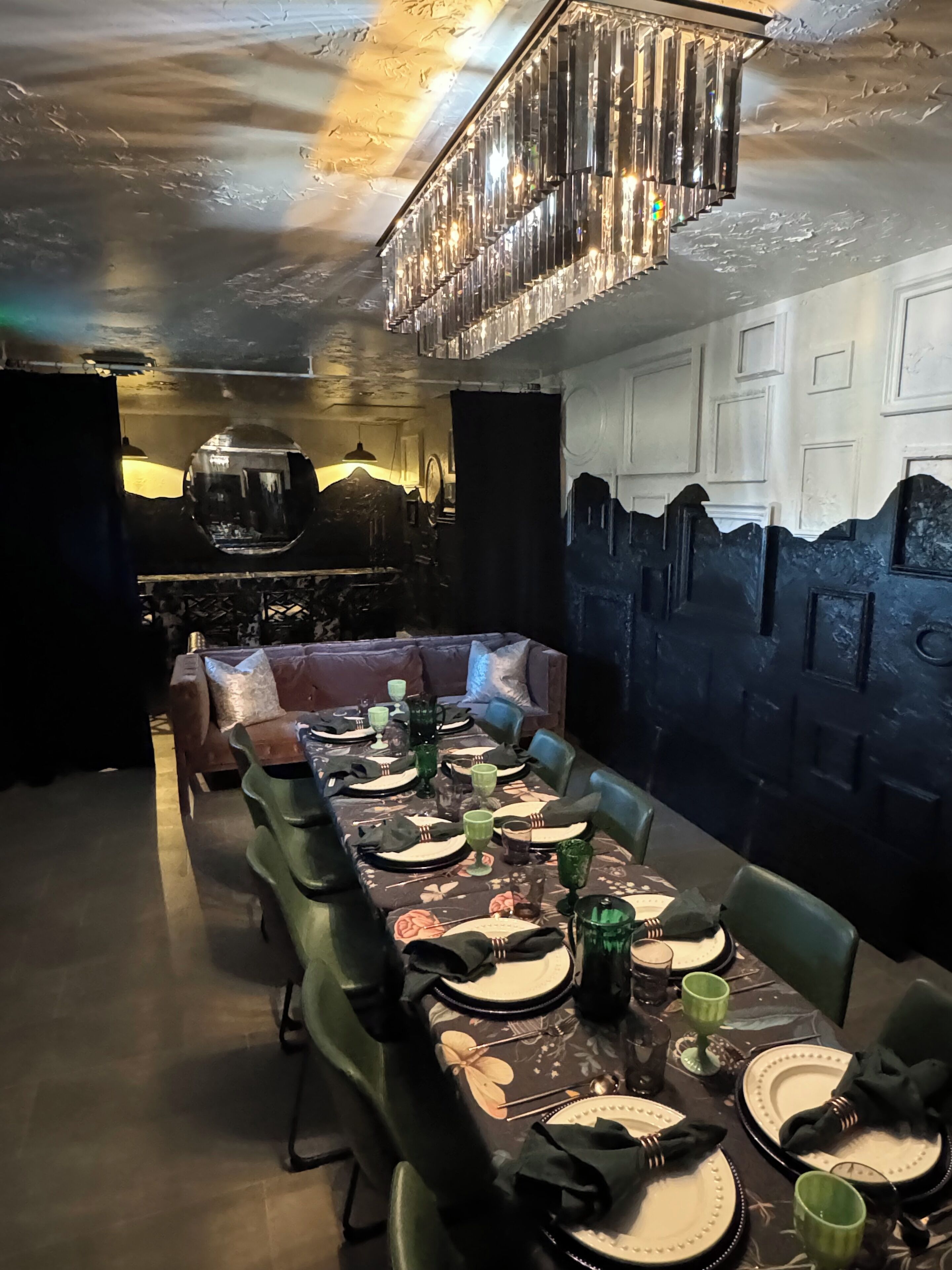 A dining area with a long table set for a meal, a chandelier, and a wall with a mountain design.