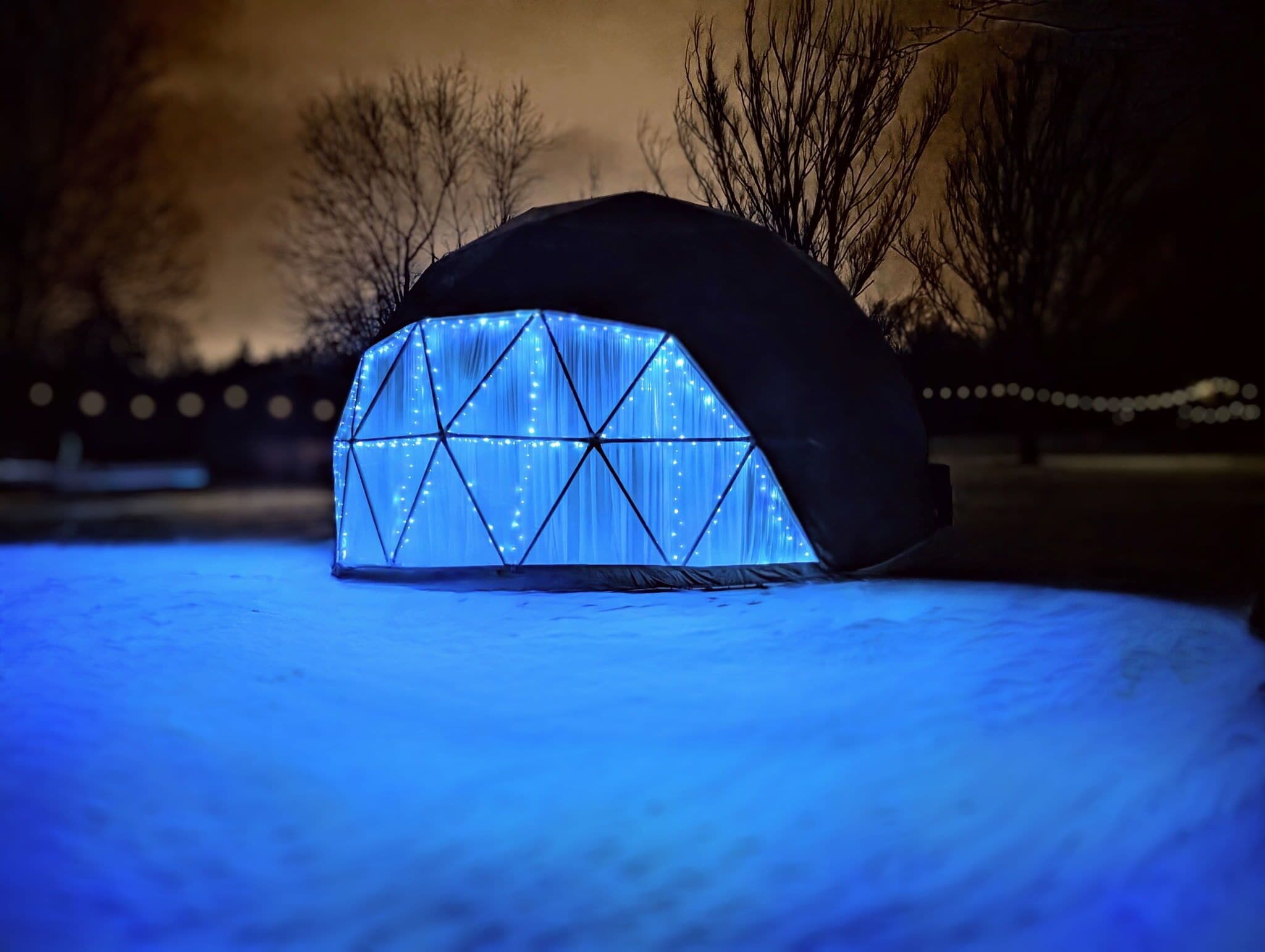 A geodesic dome structure with a grid pattern and illuminated interior, situated on snow-covered ground.