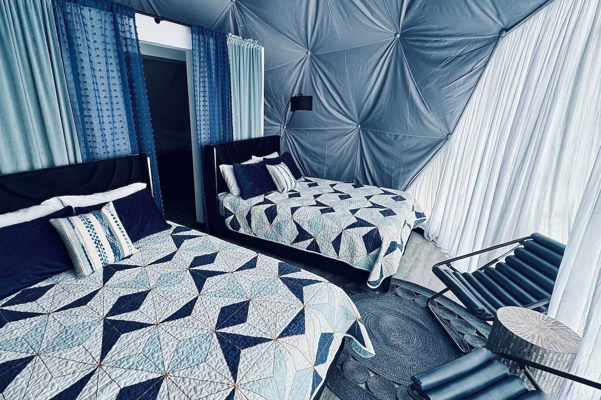 A hotel room with two beds, each with a geometric patterned bedspread, a large tufted headboard, and a chair with a woven seat.