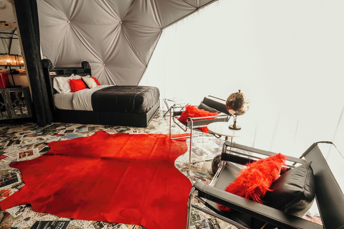 A modern bedroom with a large bed, a red rug, a chaise lounge, and a unique geometric ceiling design.